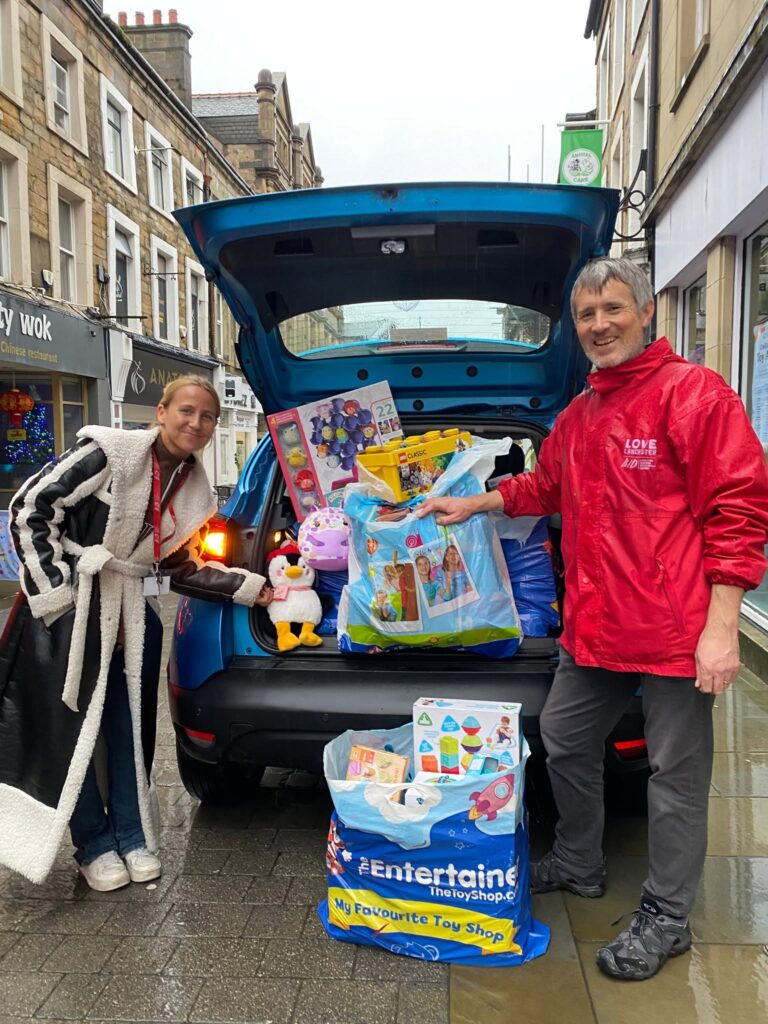 Two people stood at the boot of a blue car on a high street. The car boot is filled with toys in shopping bags. There is also a bag full of toys on the ground behind the car.